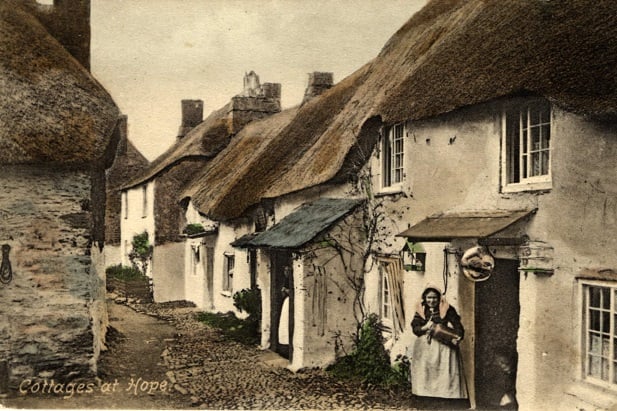 Hand-coloured picture postcard of cottages at Hope Cove. Lady holding small churn in doorway.